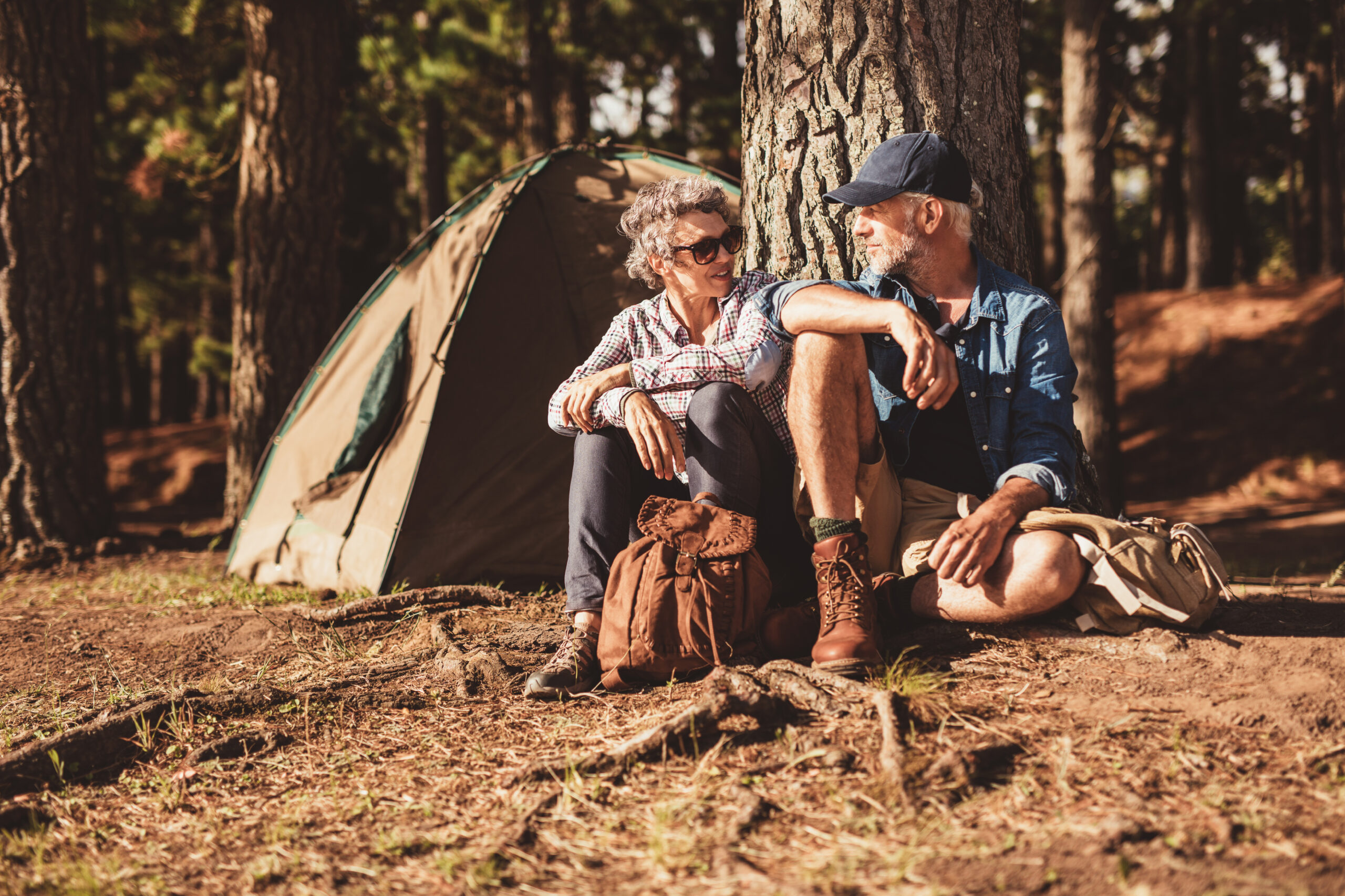 Portrait of happy senior couple sitting together under a tree on summer day. Mature man and woman near the tent, camping in forest.