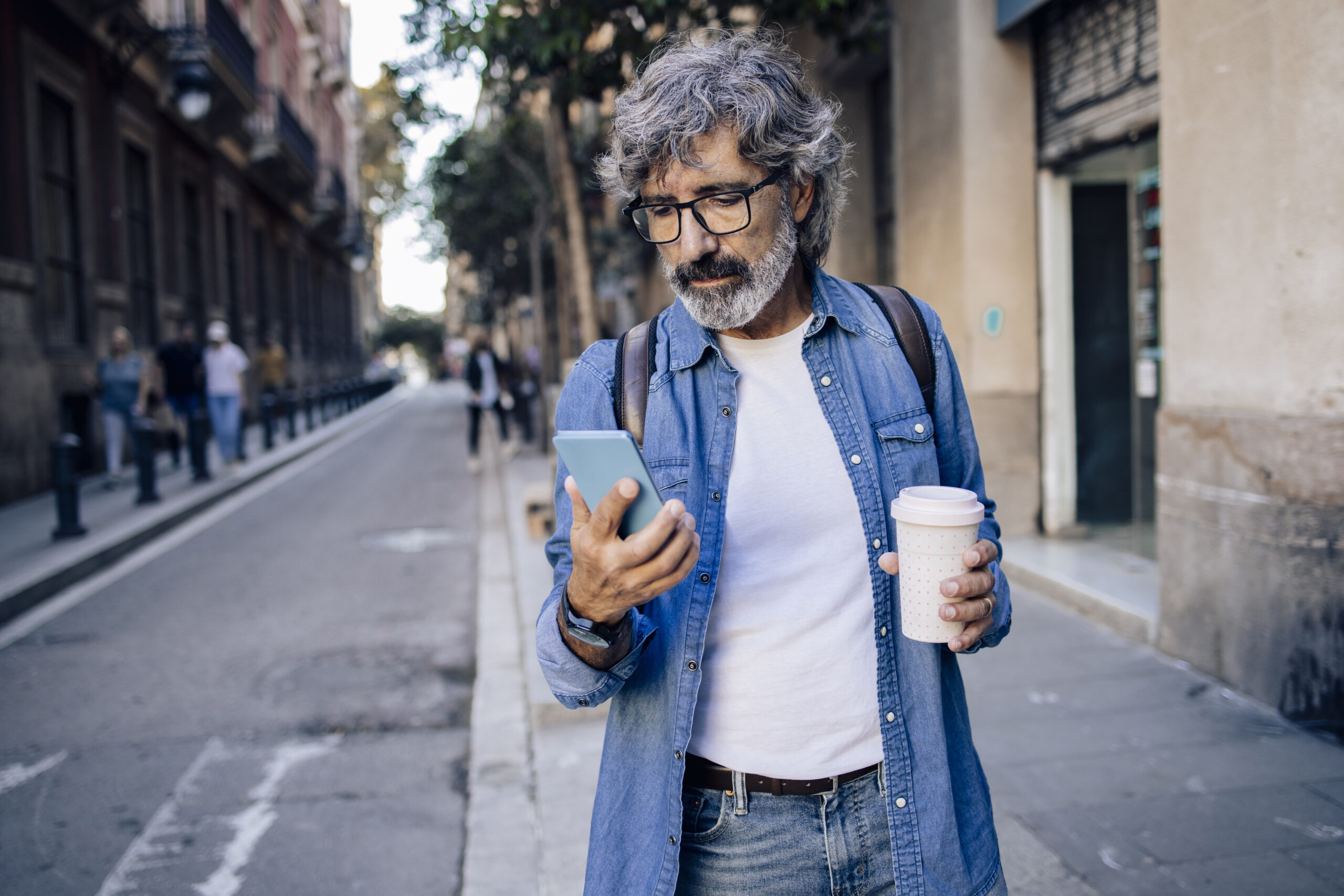 Portrait of a mature man on vacation sightseeing Barcelona, enjoying coffee and using smartphone on the move.