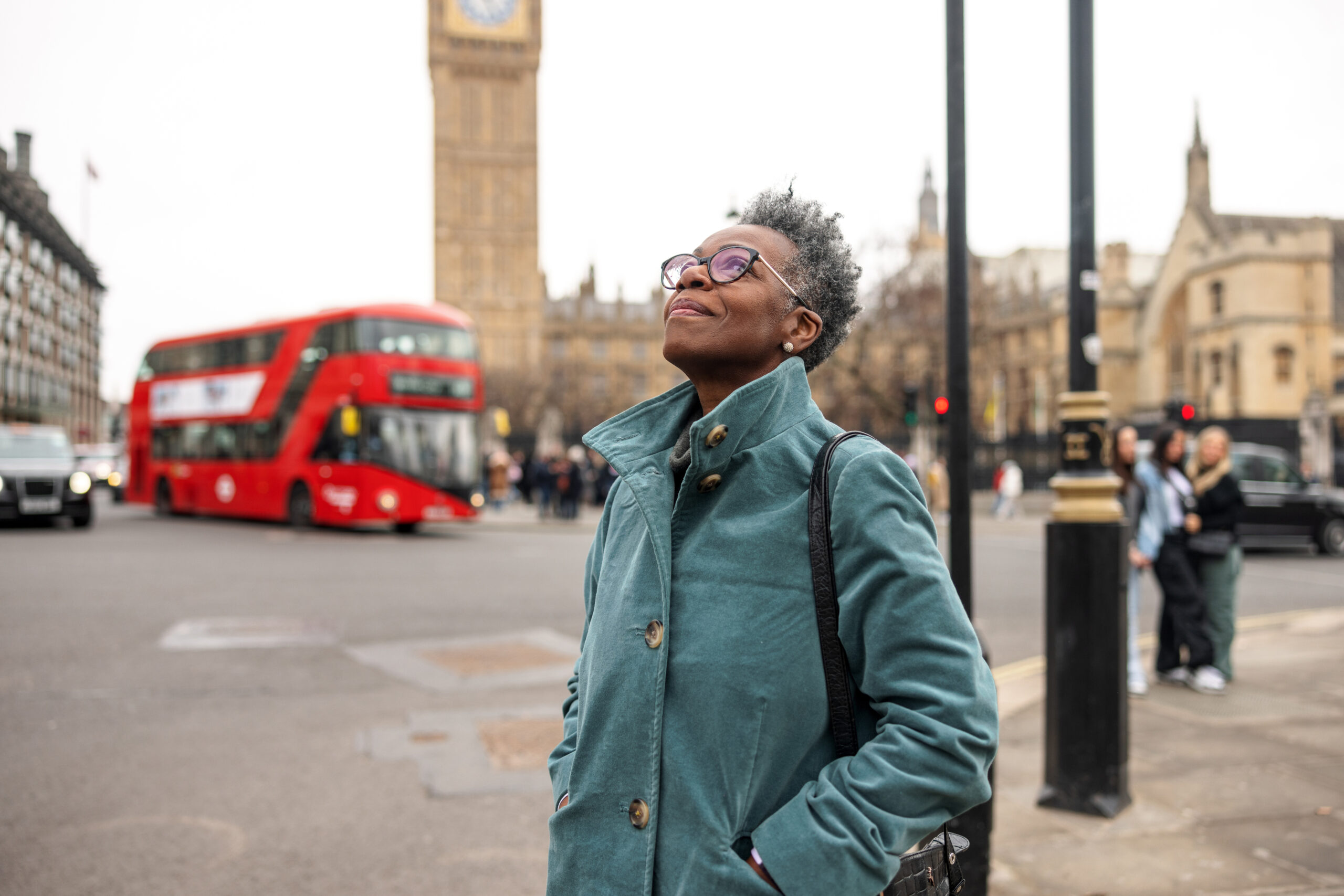 A senior adult black female tourist admiring the architecture in London. She is traveling alone and walking around the City of Westminster on a gloomy winter day. The woman is standing on the sidewalk and behind her, there is the famous Big Ben. She is looking up